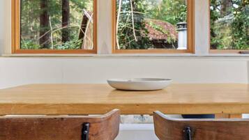A dining area with a wooden table, two chairs, and a view of a forest through large windows.
