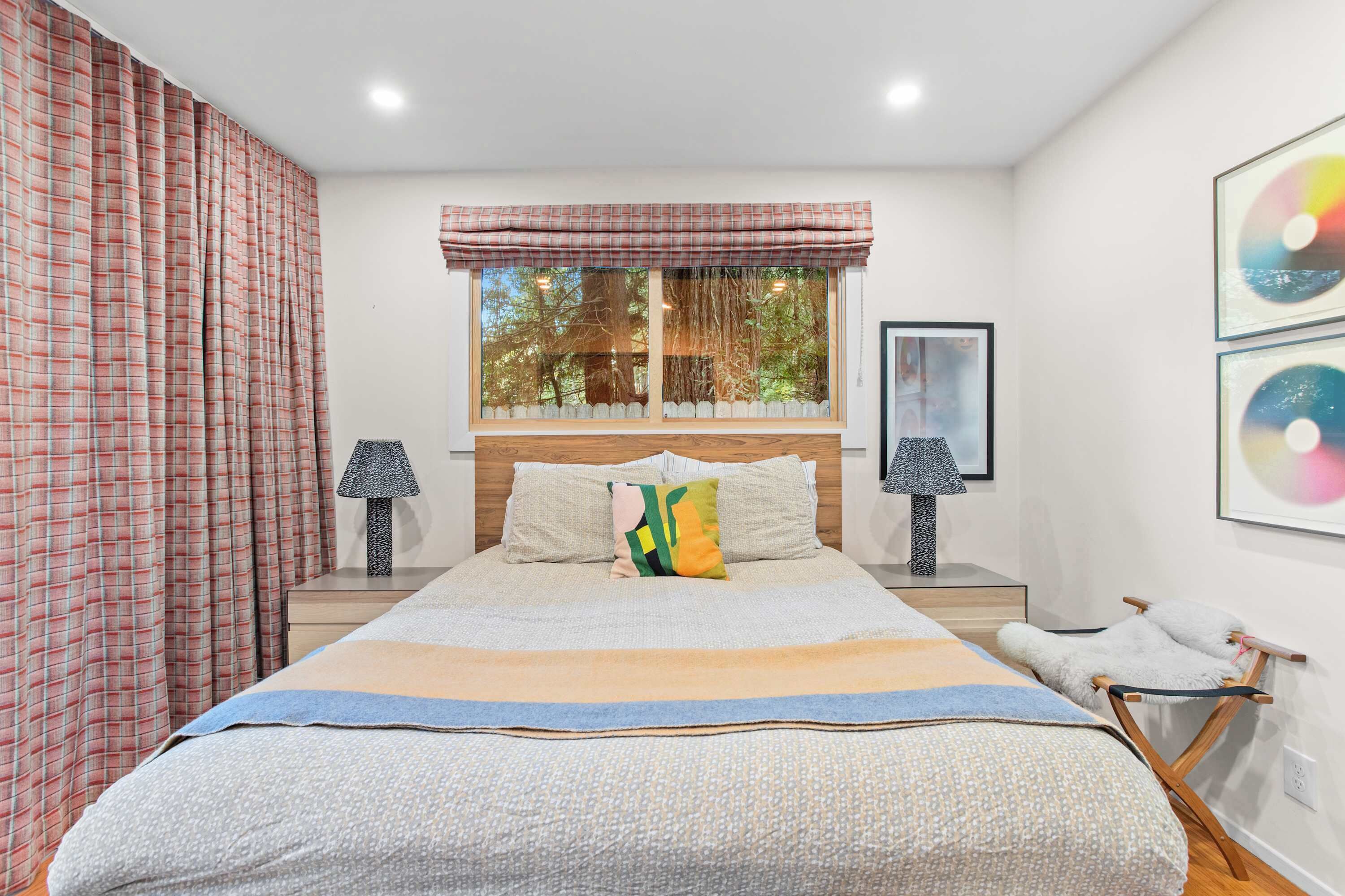 A bedroom with a large bed, two bedside tables, a window with curtains, and wall art.