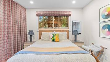A bedroom with a large bed, two bedside tables, a window with curtains, and wall art.
