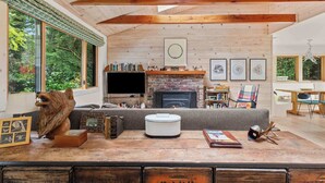 A cozy living room with a fireplace, wooden beams, a large window, and a cat statue. - Cozy Riverfront Cabin with Dock, Fire Pit & Dogs OK (Monte Rio)