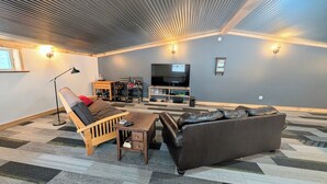 A living room with a sofa, a recliner, a coffee table, a TV, and a shelf with various items. - Barndominium in RACO, located very close to trails. (brimley)