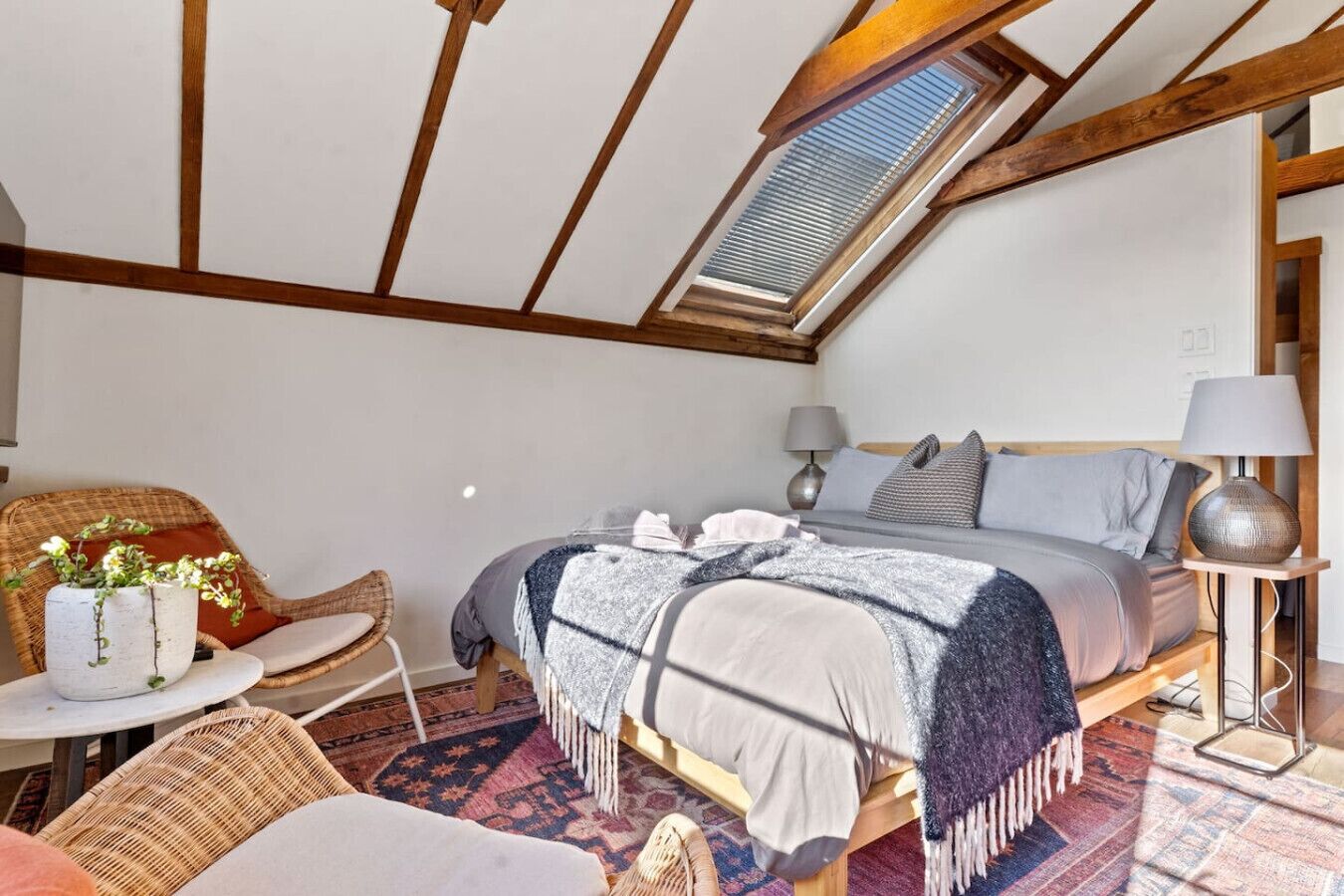 A bedroom with a sloped ceiling, a bed with a grey blanket, a side table with a lamp, and a wicker chair.
