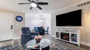 Een woonkamer met een flatscreen televisie, een open haard, een plafondventilator en een eethoek met blauwe stoelen.