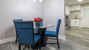 Een eetgedeelte met een ronde tafel en vier blauwe stoelen, een potplant met rode bloemen, en een moderne keuken op de achtergrond.
