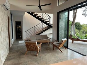 A modern interior with a staircase, a seating area with wooden furniture, and a large glass door leading to an outdoor area. - Tranquil Hillside 3-Bedroom Home with Nosara Ocean Views (Arenals de Nosara)