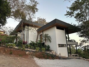 A modern two-story building with a flat roof, large windows, and a covered outdoor seating area. - Tranquil Hillside 3-Bedroom Home with Nosara Ocean Views (Arenals de Nosara)