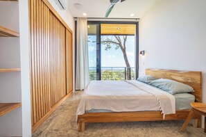 A bedroom with a wooden bed, a view of a tree through a large window, and a balcony. - Tranquil Hillside 3-Bedroom Home with Nosara Ocean Views (Arenals de Nosara)