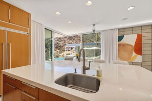A modern kitchen with a large island, a stainless steel sink, and a view of a pool and mountains. - Glass Haus - Desert Retreat with Epic Views! (Palm Springs)