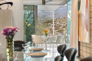 A modern dining area with a glass table and chairs, a vase with flowers, and a view of a mountainous landscape. - Glass Haus - Desert Retreat with Epic Views! (Palm Springs)