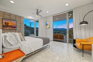 A modern bedroom with a bed, a nightstand, a chair, and a large window with a view of the landscape. - Glass Haus - Desert Retreat with Epic Views! (Palm Springs)