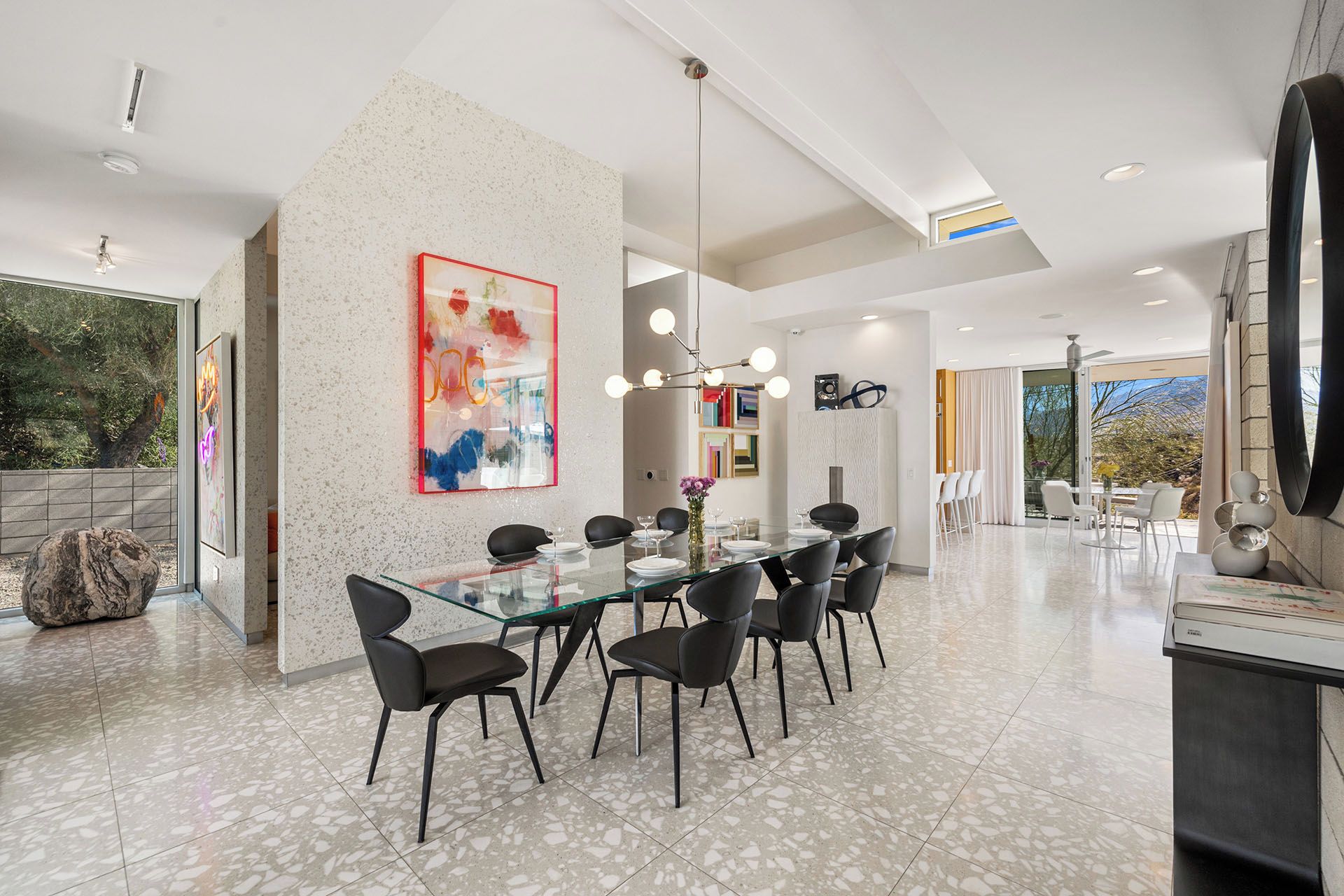 Uma ampla área de jantar com mesa de vidro e cadeiras pretas, um grande quadro abstrato na parede e vista para o exterior.