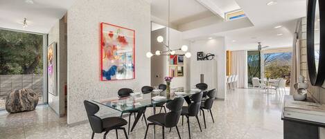 Uma ampla área de jantar com mesa de vidro e cadeiras pretas, um grande quadro abstrato na parede e vista para o exterior.