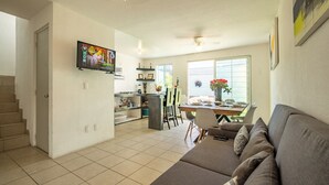 Una sala de estar moderna con un sofá gris, un comedor con mesa y sillas, una cocina con refrigerador y estantes, y un televisor montado en la pared.