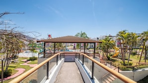 Un sendero conduce a una zona de juegos con un área de descanso cubierta y palmeras.