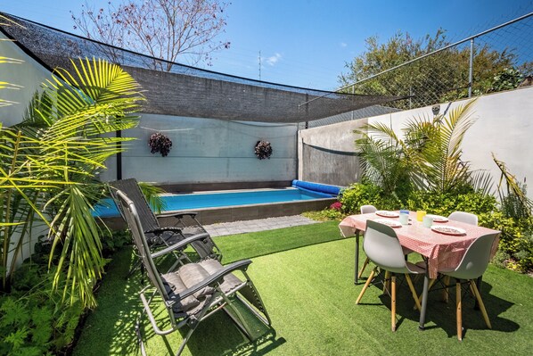 Un jardin avec une piscine, du gazon synthétique, une table à manger avec des chaises et un fauteuil de relaxation.