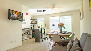 Una sala moderna con un sofá gris, un comedor con mesa y sillas, y una cocina con refrigerador y televisor montado en la pared.