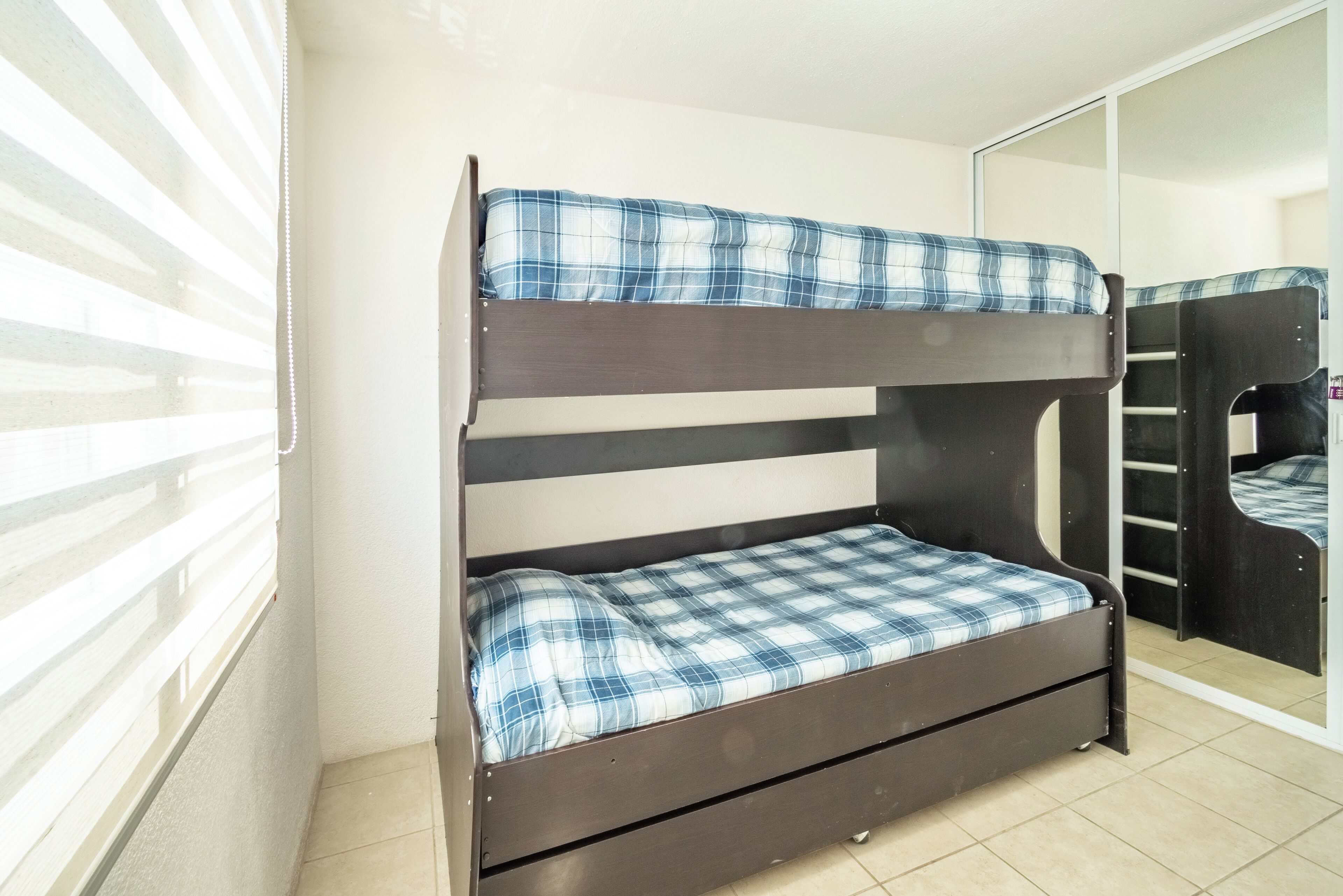 A bunk bed with a blue and white checkered bedspread, a wooden headboard, and a mirrored closet door.
