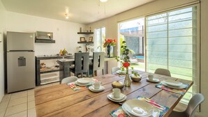 Cocina moderna con comedor, que incluye una mesa de madera dispuesta para cuatro personas, un refrigerador y una ventana con vista a vegetación.