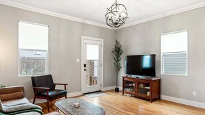 Ein Wohnzimmer mit einer Couch, einem Sessel, einem Couchtisch, einem Fernseher auf einem Holzboden und einer Tür, die auf einen Balkon führt.