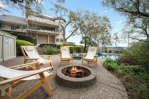 A fire pit surrounded by lounge chairs, a swimming pool, and a building with a balcony.