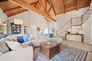 A spacious living room with a high wooden ceiling, a large beige sectional sofa, a wooden coffee table, and a staircase with wooden railings.