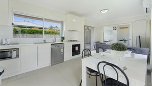 Une cuisine moderne avec des meubles blancs, un lave-vaisselle, une cuisinière et un évier. On y trouve un coin repas avec une table et des chaises blanches, et une fenêtre donnant sur une maison.