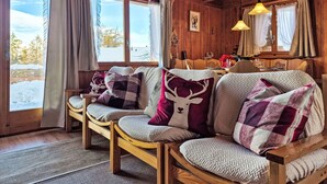 A cozy cabin interior with wooden walls and ceiling, a sofa set, a dining table, and a view of a snowy landscape through large windows. - La Tanière by Interhome (Nendaz)