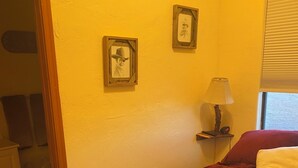 Une chambre à coucher avec une couvre-lit rouge, deux tableaux encadrés au mur, une lampe de chevet et une fenêtre avec des stores.
