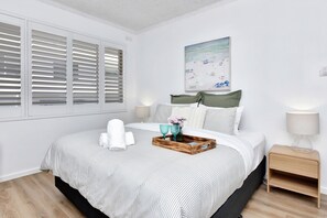 A neatly arranged bedroom with a bed, pillows, a bedside table, a lamp, and a wall-mounted painting. - Del Rae 14 Shoal Bay Escape (Shoal Bay)