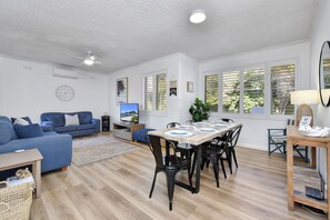 A modern living room with a blue sofa, a dining table with chairs, and a TV. - Del Rae 14 Shoal Bay Escape (Shoal Bay)