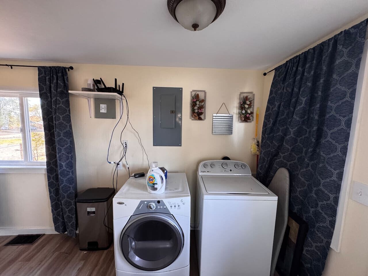 Ein Waschraum mit einer Waschmaschine und einem Trockner mit Frontlader, einem kleinen Mülleimer, einer Steckdose und einem Fenster mit Vorhängen.