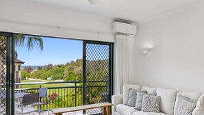 Una sala de estar con un sofá blanco, una mesita redonda y vistas a un paisaje verde a través de una puerta corrediza de vidrio.