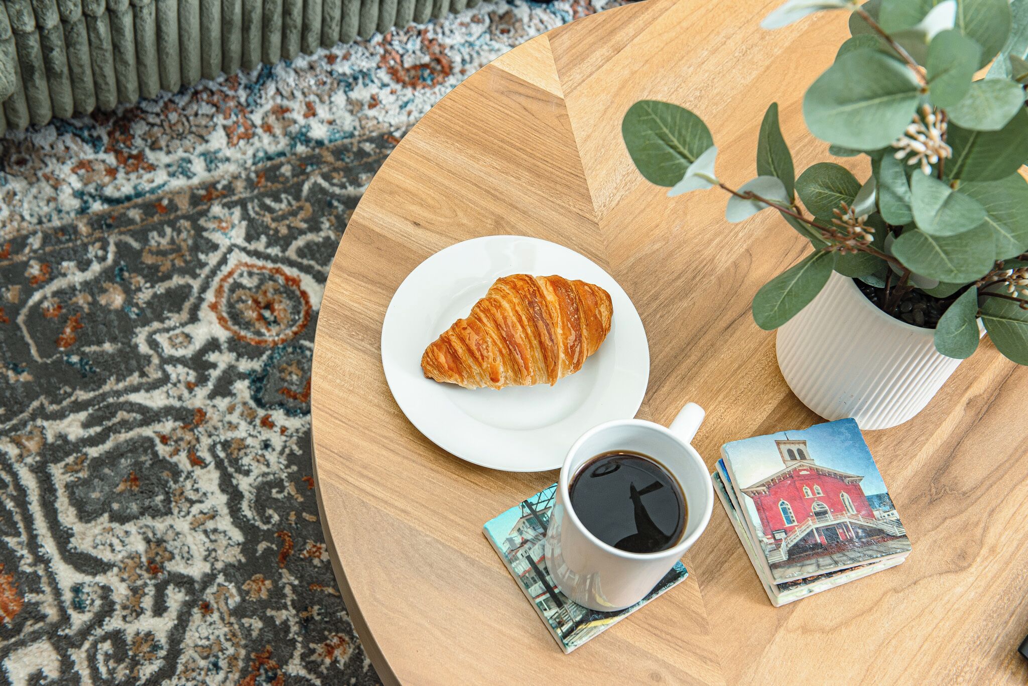A round wooden table with a croissant on a white plate, a cup of coffee, and postcards.