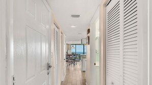 A hallway with white doors, a tiled floor, and a view of a dining area with a table and chairs. - Beautiful Condo with Panoramic Ocean Views: Double‑Wide Balcony, Heated Pool & Hot Tub ☀️ (Cape Canaveral)