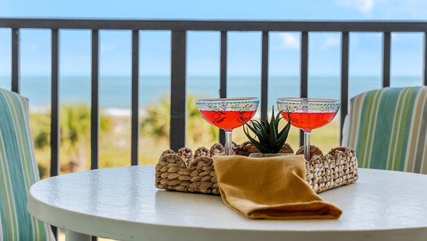A round table with two drinks, a woven basket, and a small plant on a balcony overlooking the ocean. - Beautiful Condo with Panoramic Ocean Views: Double‑Wide Balcony, Heated Pool & Hot Tub ☀️ (Cape Canaveral)