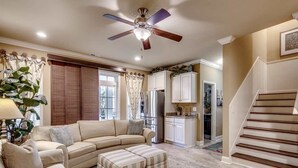 A living room with a sectional sofa, ottoman, ceiling fan, kitchen area, and a staircase. - Landyn Beach House (Myrtle Beach)