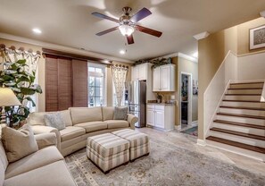 A living room with a sectional sofa, ottoman, ceiling fan, kitchen area, and a staircase. - Landyn Beach House (Myrtle Beach)