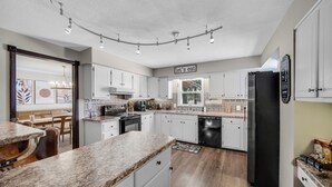 Una cocina moderna con armarios blancos, encimeras de granito y un refrigerador negro. Hay una zona de comedor con sillas de madera y una mesa.