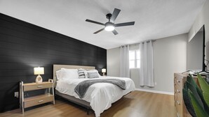 Un dormitorio con cama, mesitas de noche, ventilador de techo y ventana con cortinas.