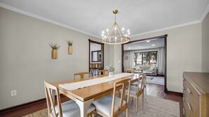 Un comedor con una mesa de madera, sillas, una araña y vistas a una sala de estar.