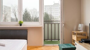 Une chambre à coucher avec un lit, un petit bureau avec un ordinateur, une fenêtre donnant sur des immeubles et un radiateur.