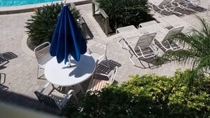 En pool med solstolar och parasoller, omgiven av palmer och en byggnad i bakgrunden.