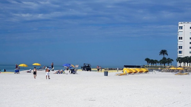 En strand med vit sand, gula parasoller och några få personer. I bakgrunden syns en byggnad omgiven av palmer.