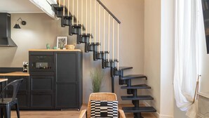Una cocina moderna con una escalera negra, una zona de comedor con una silla de mimbre y una pequeña mesa con cojín blanco y negro.