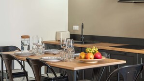 Un comedor con una mesa preparada para dos personas, un frutero y una cocina al fondo.