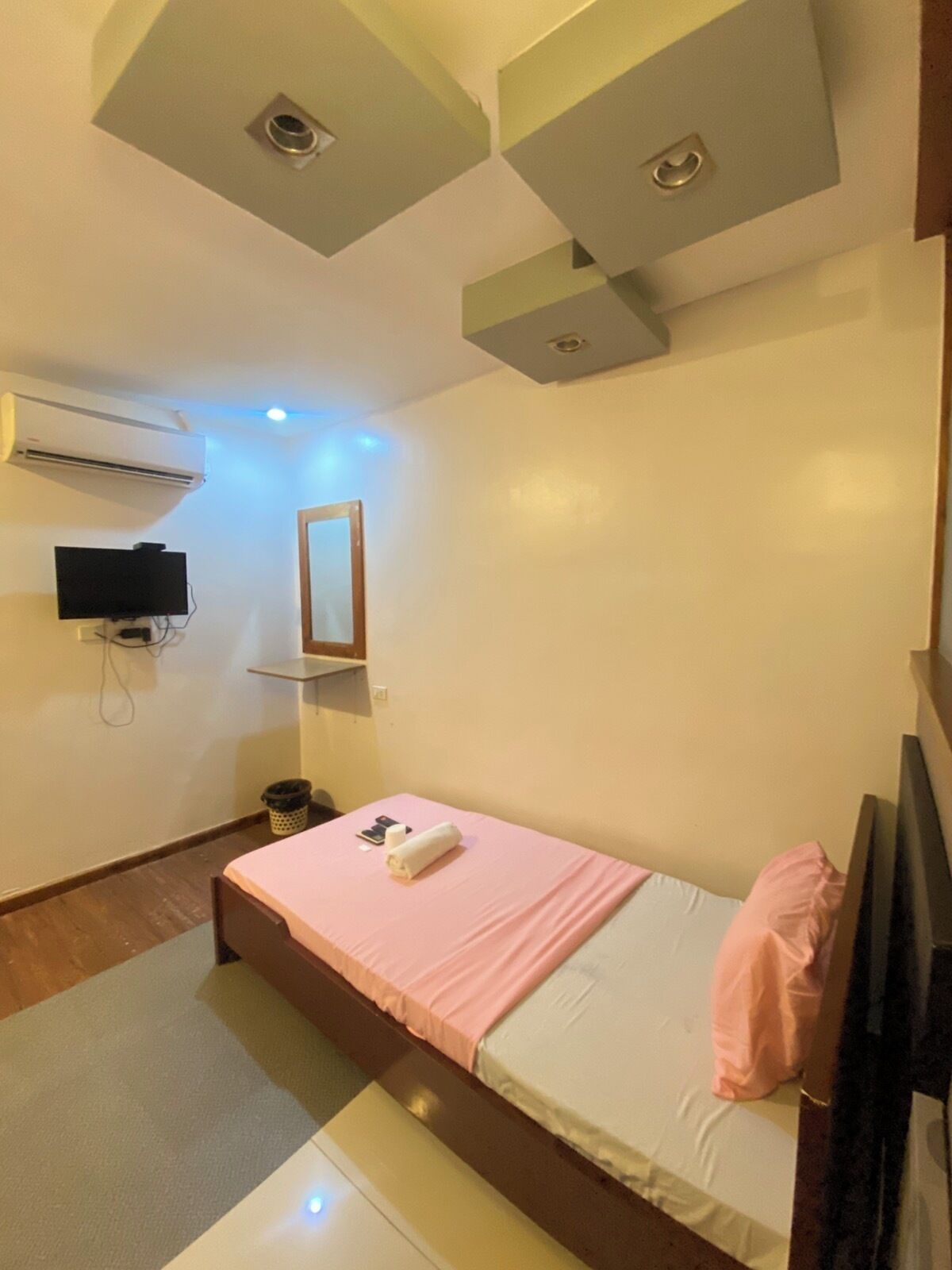 Quarto com uma cama de solteiro, TV de tela plana na parede, ar-condicionado e iluminação embutida no teto.