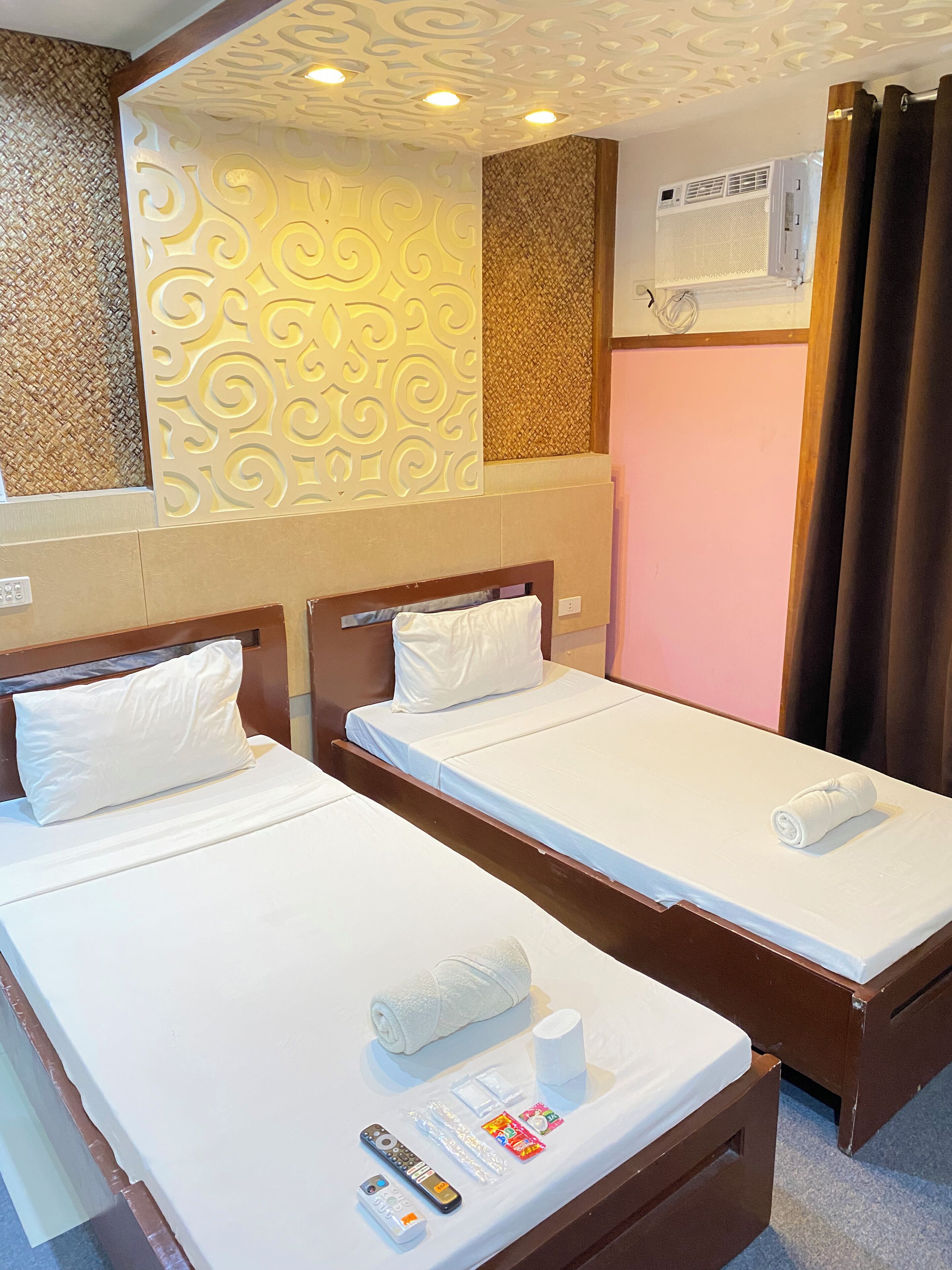 A hotel room with two single beds, a wall-mounted air conditioner, and a decorative wall panel.