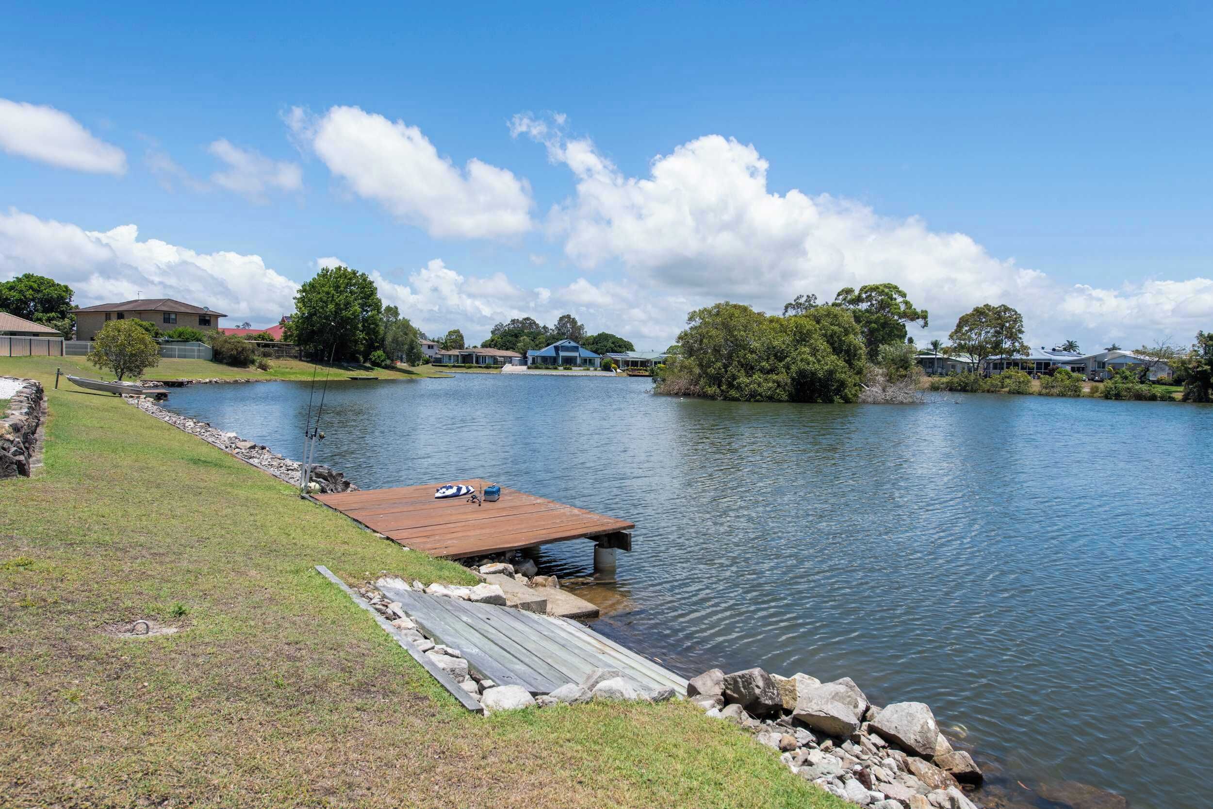 A lakeside property with a wooden dock extending into the water, surrounded by grassy areas and residential buildings in the background.