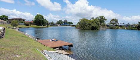 A lakeside property with a wooden dock extending into the water, surrounded by grassy areas and residential buildings in the background.