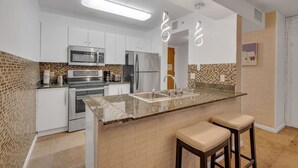 A modern kitchen with white cabinets, stainless steel appliances, a granite countertop, and two bar stools. - Amazing  unit/location with amenities & parking (Miami)